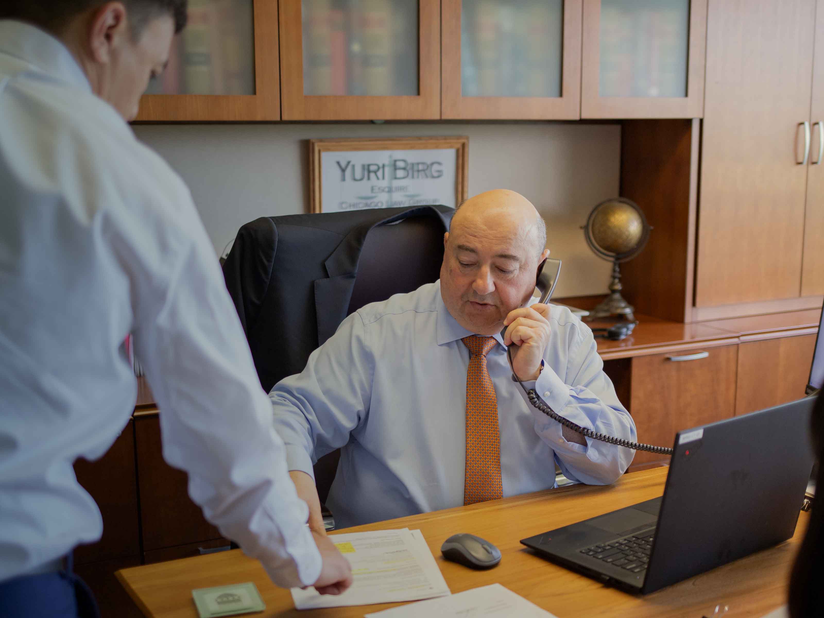Attorney Yuri Birg takes a client call at his office desk while a colleague hands him case documents